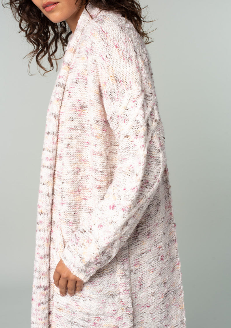 [Color: Natural/Berry] A half body side facing image of a brunette model wearing a natural off white and berry speckled knit long cardigan with an open front, side pockets, and cable knit details. 