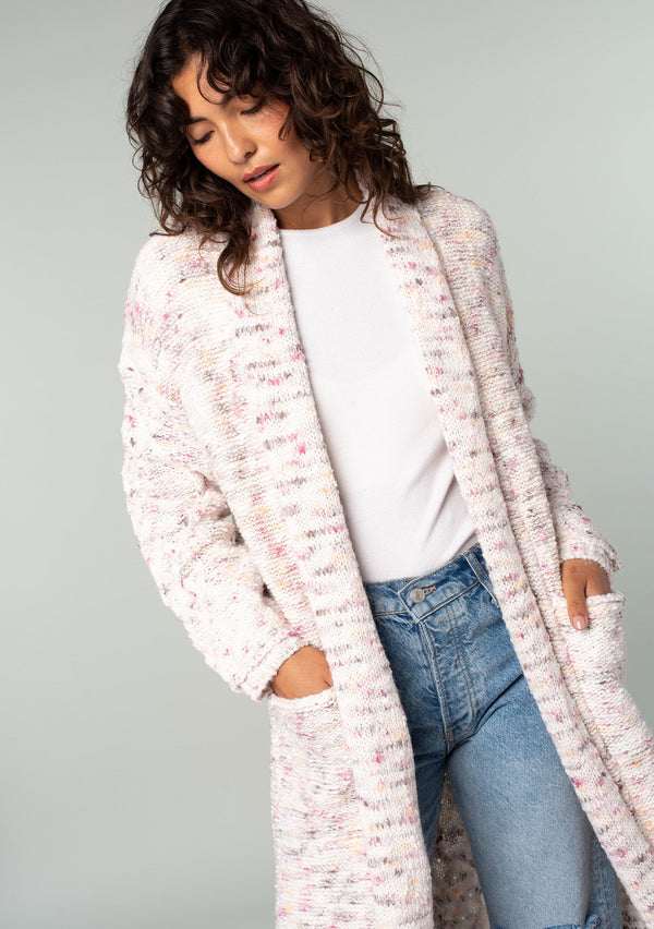 [Color: Natural/Berry] A half body front facing image of a brunette model wearing a natural off white and berry speckled knit long cardigan with an open front, side pockets, and cable knit details. 