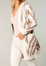 [Color: Ivory/Rust] A model wearing an ivory and rust tiger print cardigan. Featuring a mid length, long sleeves, side pockets, and an open front.