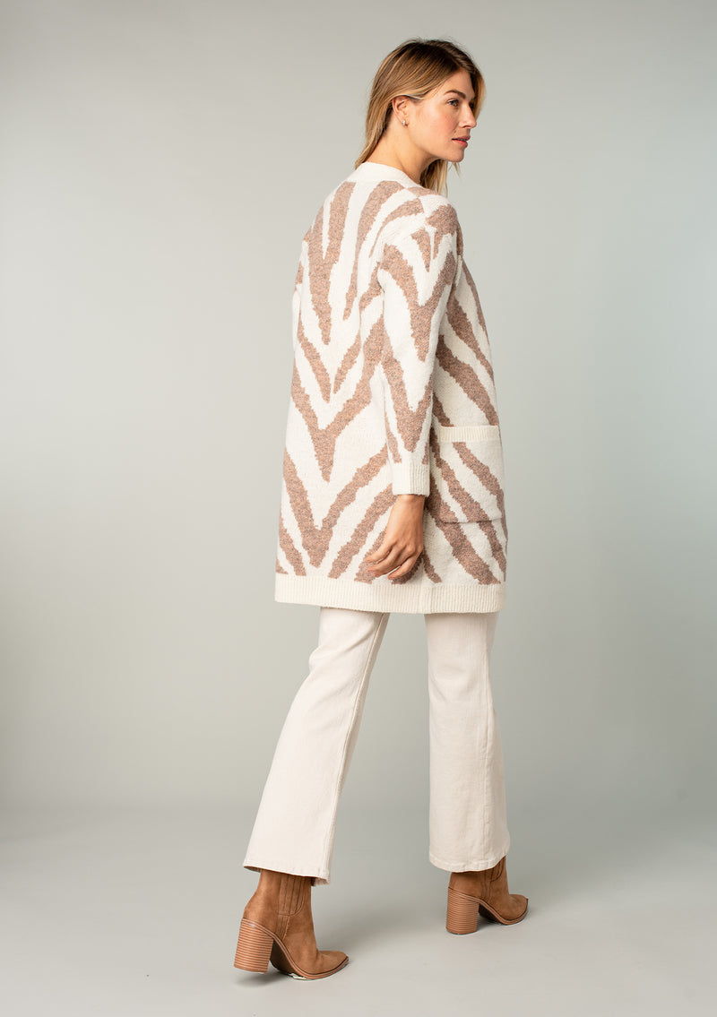 [Color: Ivory/Rust] A model wearing an ivory and rust tiger print cardigan. Featuring a mid length, long sleeves, side pockets, and an open front.