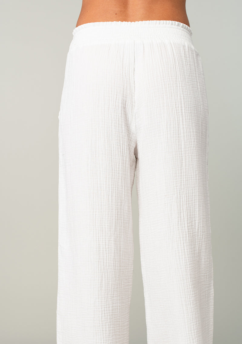 [Color: White] A back facing image of a blonde model wearing a white resort lounge pant in cotton gauze. With a wide leg, side pockets, and a smocked elastic waistband.