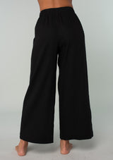 [Color: Black] A back facing image of a brunette model wearing a cool black linen and cotton blend lounge pant. With side pockets, an elastic waistband, and a wide leg. 
