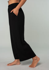 [Color: Black] A side facing image of a brunette model wearing a cool black linen and cotton blend lounge pant. With side pockets, an elastic waistband, and a wide leg. 