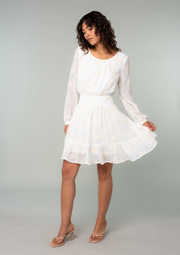 [Color: Ivory] A full body front facing image of a brunette model wearing an ivory white embroidered chiffon mini dress. Perfect for the holidays or weddings, featuring long sleeves, a tiered skirt, an open back keyhole with tie closure, and a smocked elastic waist.