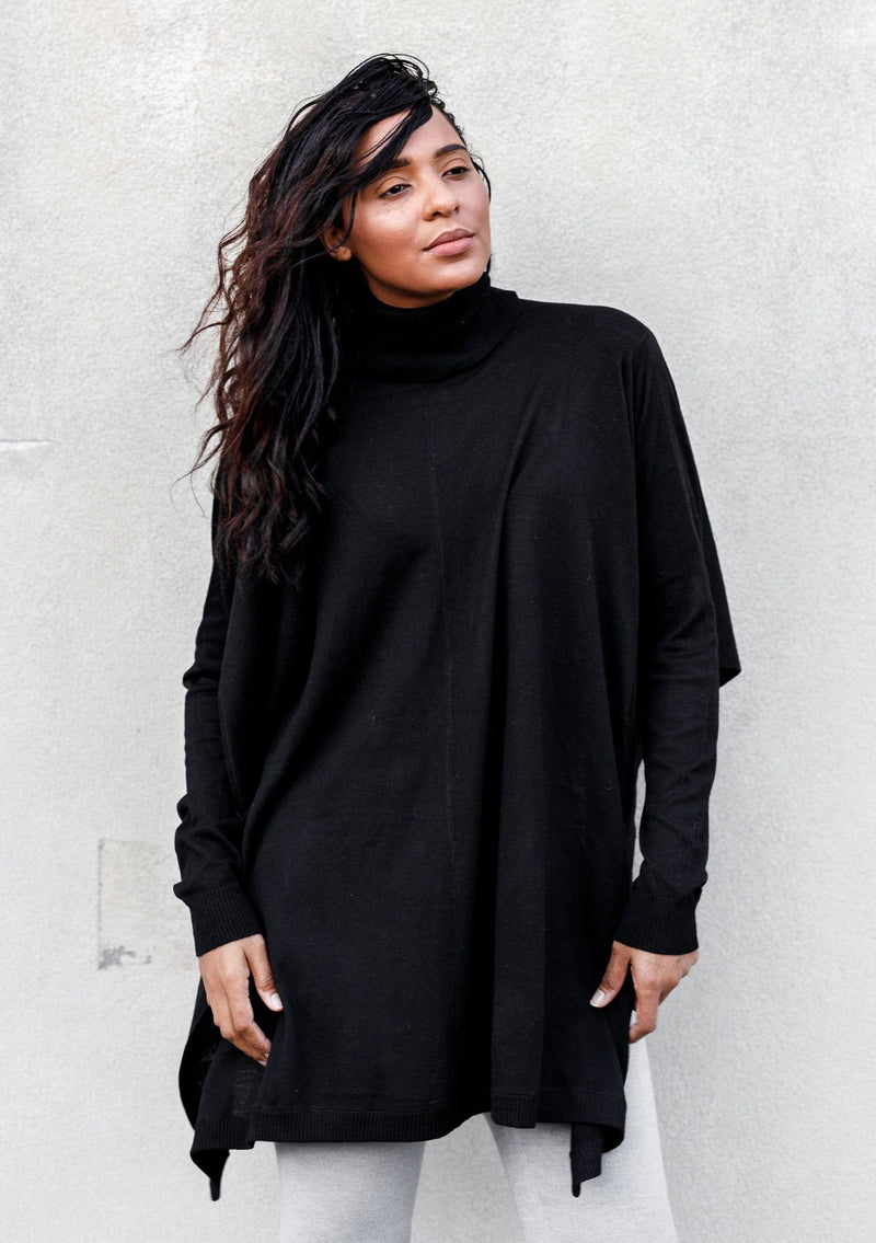 [Color: Black] A woman standing outside wearing a turtleneck sweater poncho. Featuring a flattering draped silhouette, breezy side vents, and a slightly longer tunic style length.