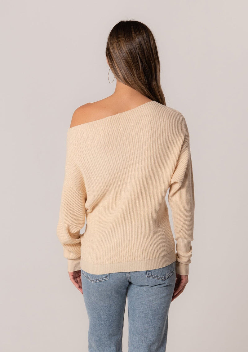 [Color: Cream] A back facing image of a brunette model wearing a cream waffle knit pullover sweater. With long sleeves, a relaxed fit, and a wide neckline that can be worn off the shoulder.