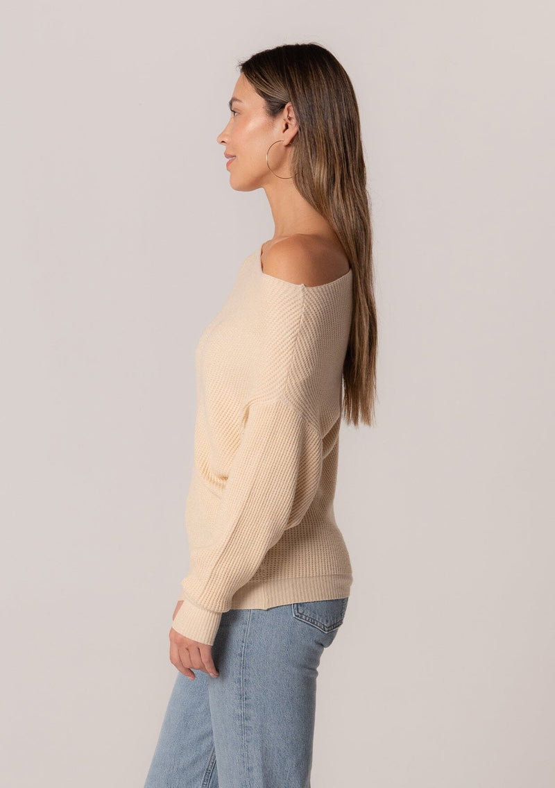 [Color: Cream] A side facing image of a brunette model wearing a cream waffle knit pullover sweater. With long sleeves, a relaxed fit, and a wide neckline that can be worn off the shoulder.