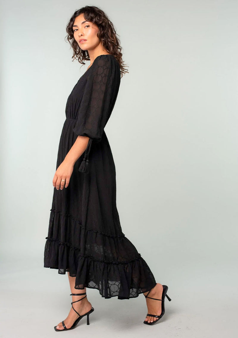 [Color: Black] A front facing image of a brunette model wearing a bohemian black maxi dress in embroidered chiffon. With three quarter length sleeves, adjustable tie cuffs, a self covered button front, an elastic waist, and a ruffle trimmed tiered skirt.