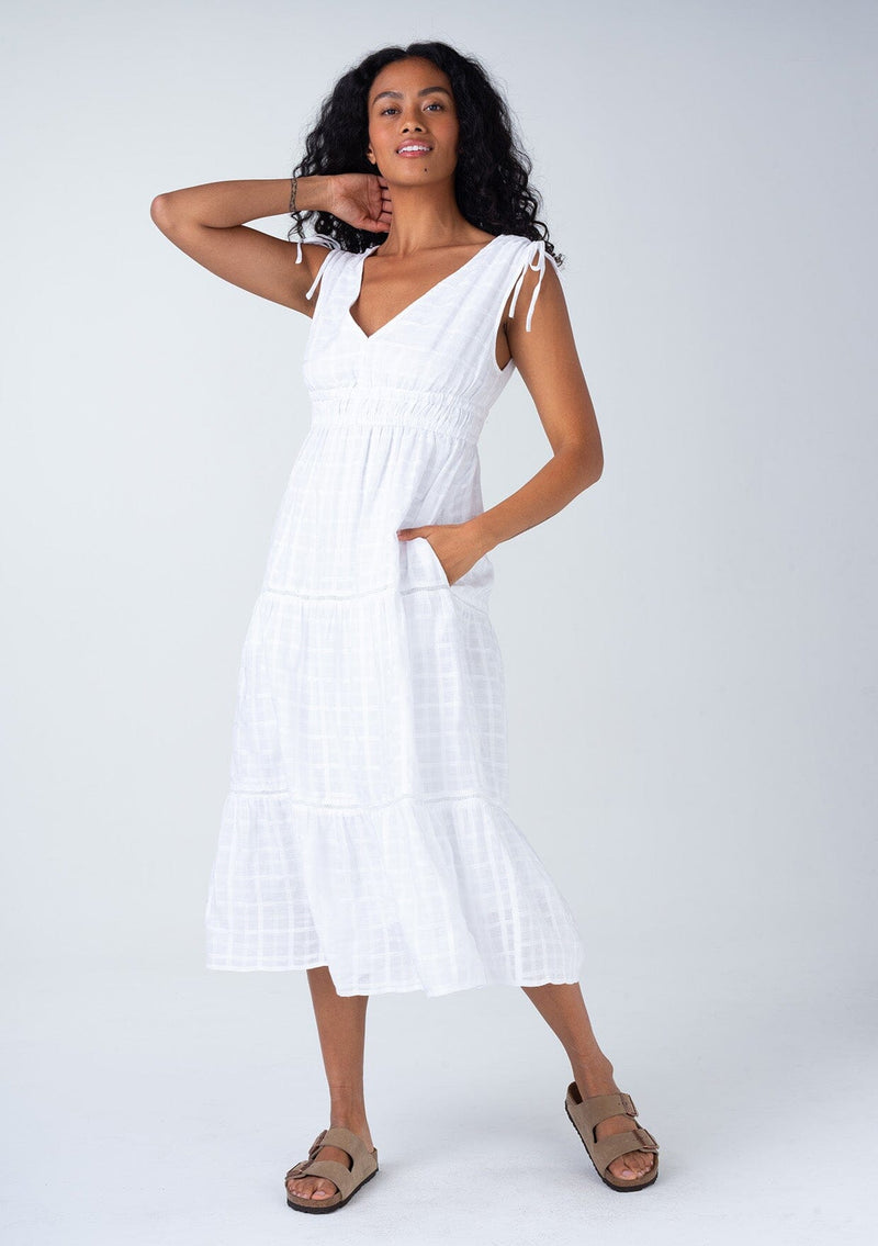 [Color: White] A front facing image of a brunette model wearing a classic bohemian spring mid length dress in a white cotton textured gingham. With adjustable shoulder ties, a v neckline in front and back, a lace trimmed tiered skirt, an elastic waist, and side pockets. 