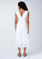 [Color: White] A back facing image of a brunette model wearing a classic bohemian spring mid length dress in a white cotton textured gingham. With adjustable shoulder ties, a v neckline in front and back, a lace trimmed tiered skirt, an elastic waist, and side pockets. 