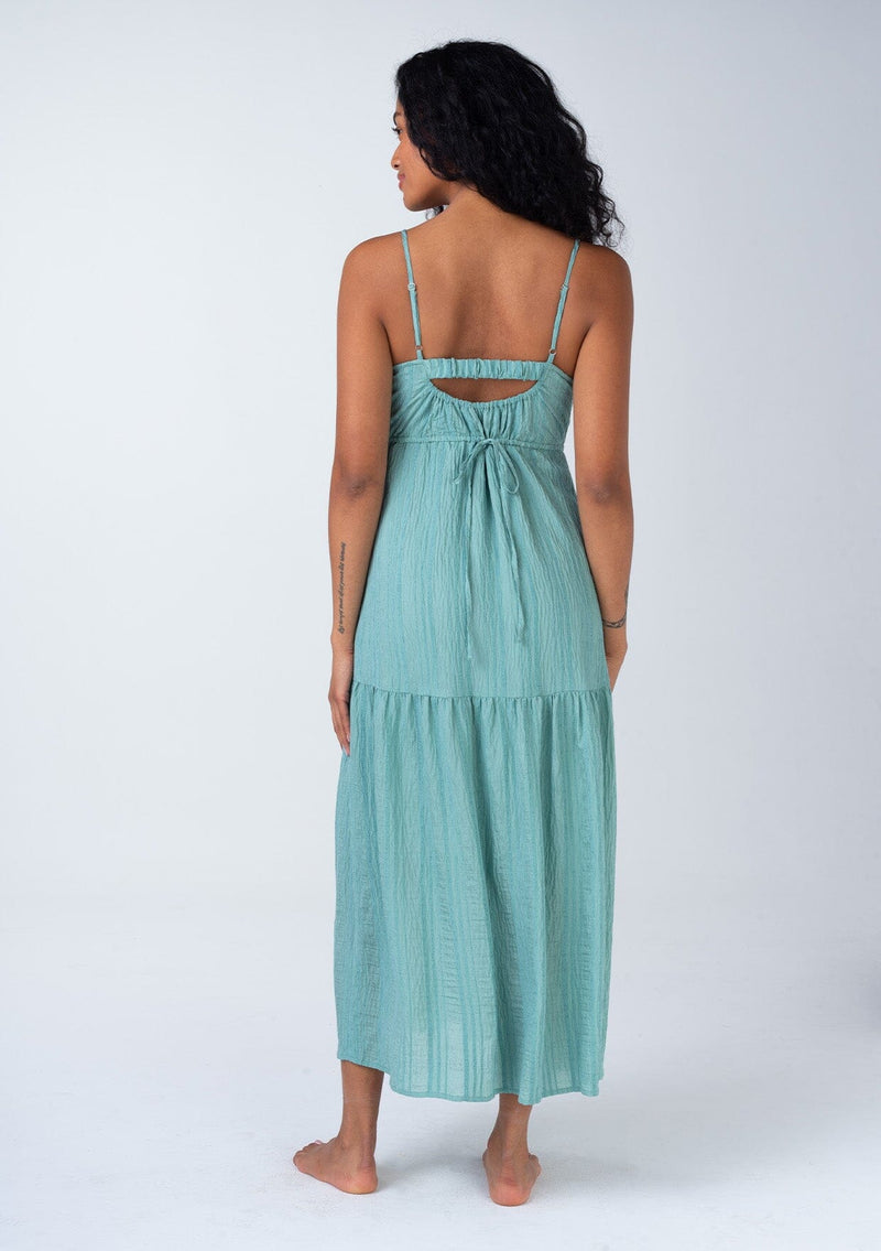 [Color: Dusty Teal] A back facing image of a brunette model wearing a bohemian spring maxi tank dress in a dusty teal shadow stripe. With adjustable spaghetti straps, a round neckline, a tiered flowy skirt, a back cutout detail, and a back tie waist detail.