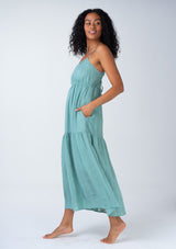 [Color: Dusty Teal] A side facing image of a brunette model wearing a bohemian spring maxi tank dress in a dusty teal shadow stripe. With adjustable spaghetti straps, a round neckline, a tiered flowy skirt, a back cutout detail, and a back tie waist detail.