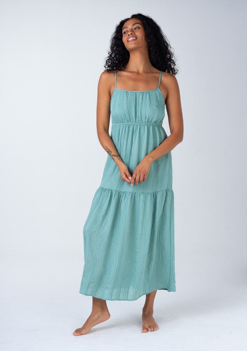 [Color: Dusty Teal] A full body front facing image of a brunette model wearing a bohemian spring maxi tank dress in a dusty teal shadow stripe. With adjustable spaghetti straps, a round neckline, a tiered flowy skirt, a back cutout detail, and a back tie waist detail.