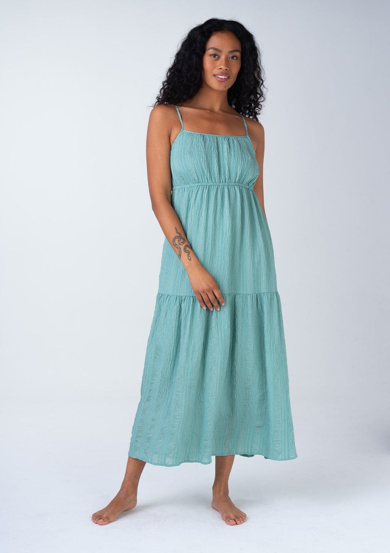 [Color: Dusty Teal] A front facing image of a brunette model wearing a bohemian spring maxi tank dress in a dusty teal shadow stripe. With adjustable spaghetti straps, a round neckline, a tiered flowy skirt, a back cutout detail, and a back tie waist detail.