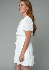 [Color: Off White] A side facing image of a red headed model wearing a white eyelet mini dress with short flutter sleeves, a split v neckline with tassel ties, and an elastic waist.