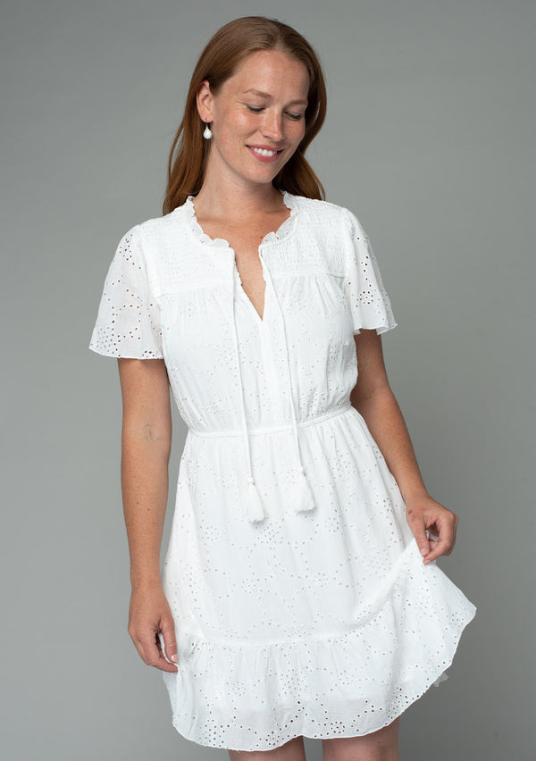 [Color: Off White] A front facing image of a red headed model wearing a white eyelet mini dress with short flutter sleeves, a split v neckline with tassel ties, and an elastic waist.