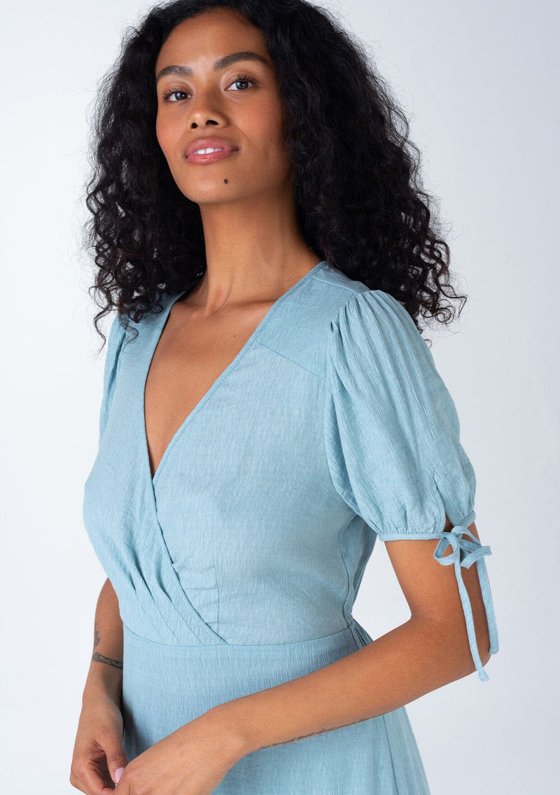 [Color: Dusty Blue] A close up front facing image of a bohemian dusty blue mid length dress. With short puff sleeves, adjustable tie cuffs, a flowy tiered skirt, and a surplice v neckline. 
