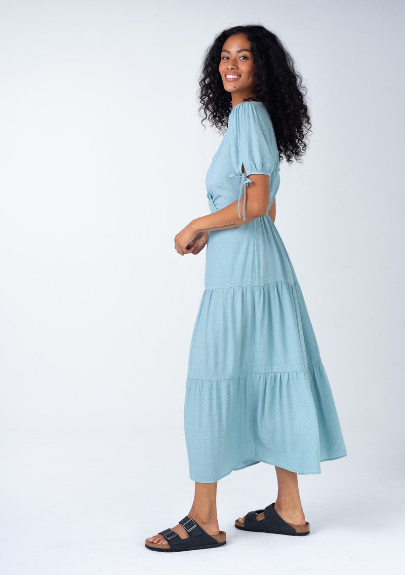 [Color: Dusty Blue] A side facing image of a bohemian dusty blue mid length dress. With short puff sleeves, adjustable tie cuffs, a flowy tiered skirt, and a surplice v neckline. 