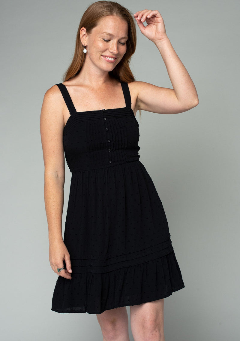 [Color: Black] A front facing image of a red headed model wearing a black sleeveless bohemian mini dress with a button front top, tank top straps, a square neckline, pleated pintuck details, and a flowy mini skirt.