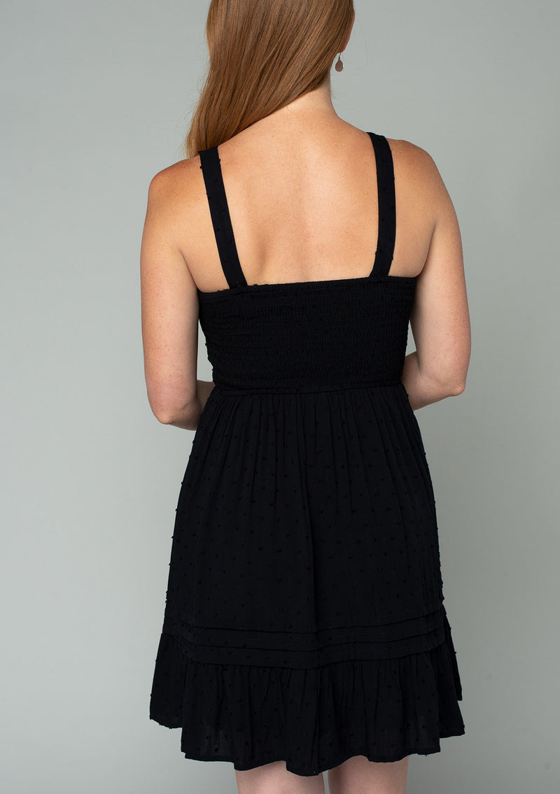 [Color: Black] A back facing image of a red headed model wearing a black sleeveless bohemian mini dress with a button front top, tank top straps, a square neckline, pleated pintuck details, and a flowy mini skirt.