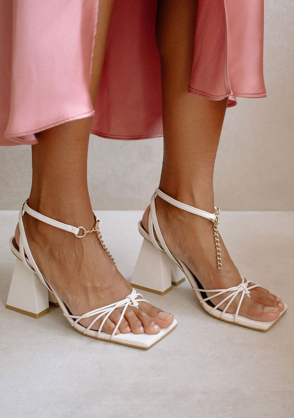 [Color: Off White] Alohas chic and trendy white leather strap block heeled sandal. With a delicate front chain accent and pyramid shaped heel with notched cutout. 
