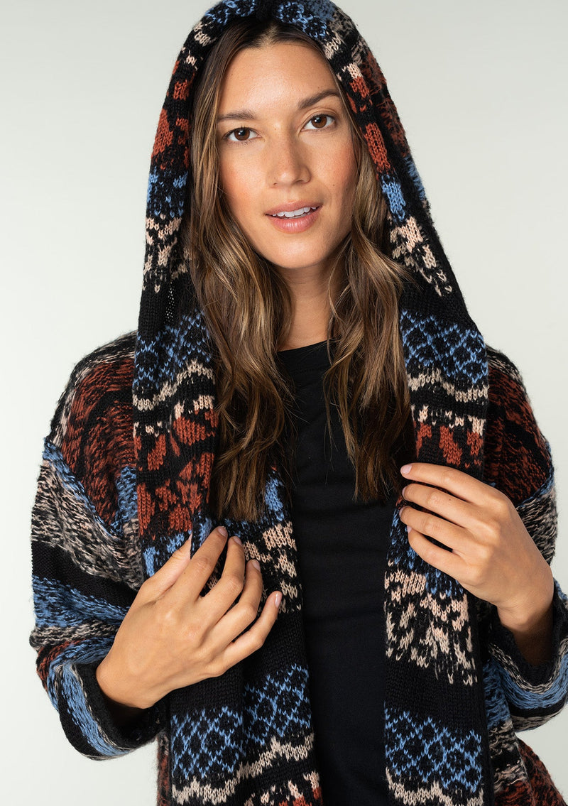 [Color: Blue/Rust] A close up front facing image of a brunette model wearing a black and rust red striped bohemian knit sweater with long sleeves, an open front, side pockets, and a warm hoodie. 