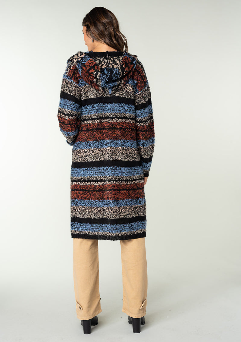 [Color: Blue/Rust] A back facing image of a brunette model wearing a black and rust red striped bohemian knit sweater with long sleeves, an open front, side pockets, and a warm hoodie. 