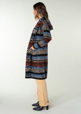 [Color: Blue/Rust] A side facing image of a brunette model wearing a black and rust red striped bohemian knit sweater with long sleeves, an open front, side pockets, and a warm hoodie. 