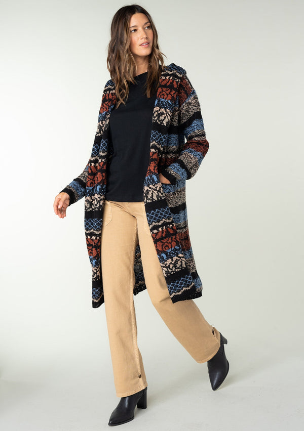 [Color: Blue/Rust] A front facing image of a brunette model wearing a black and rust red striped bohemian knit sweater with long sleeves, an open front, side pockets, and a warm hoodie. 