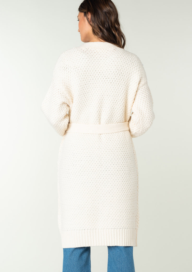[Color: Natural] A back facing image of a brunette model wearing a natural mid length cardigan in a textured honeycomb knit. With long sleeves, a tie front waist belt, and side pockets.