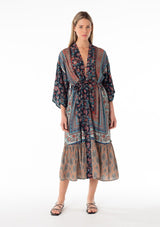 [Color: Teal/Navy] A front facing image of a blonde model wearing a bohemian mid length duster lounge robe in a mixed blue floral print. With half length long sleeves, a tiered ruffle trimmed hemline, an open front, and a tie waist belt. 