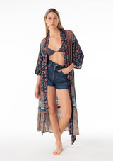 [Color: Teal/Navy] A full body front facing image of a blonde model wearing a bohemian mid length duster lounge robe in a mixed blue floral print. With half length long sleeves, a tiered ruffle trimmed hemline, an open front, and a tie waist belt. 