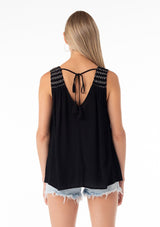 [Color: Black/Ivory] A back facing image of a blonde model wearing a bohemian black tank top. With a v neckline in the front and back, a tassel tie closure at the back, a smocked yoke detail, and contrast embroidered stitch detail. 