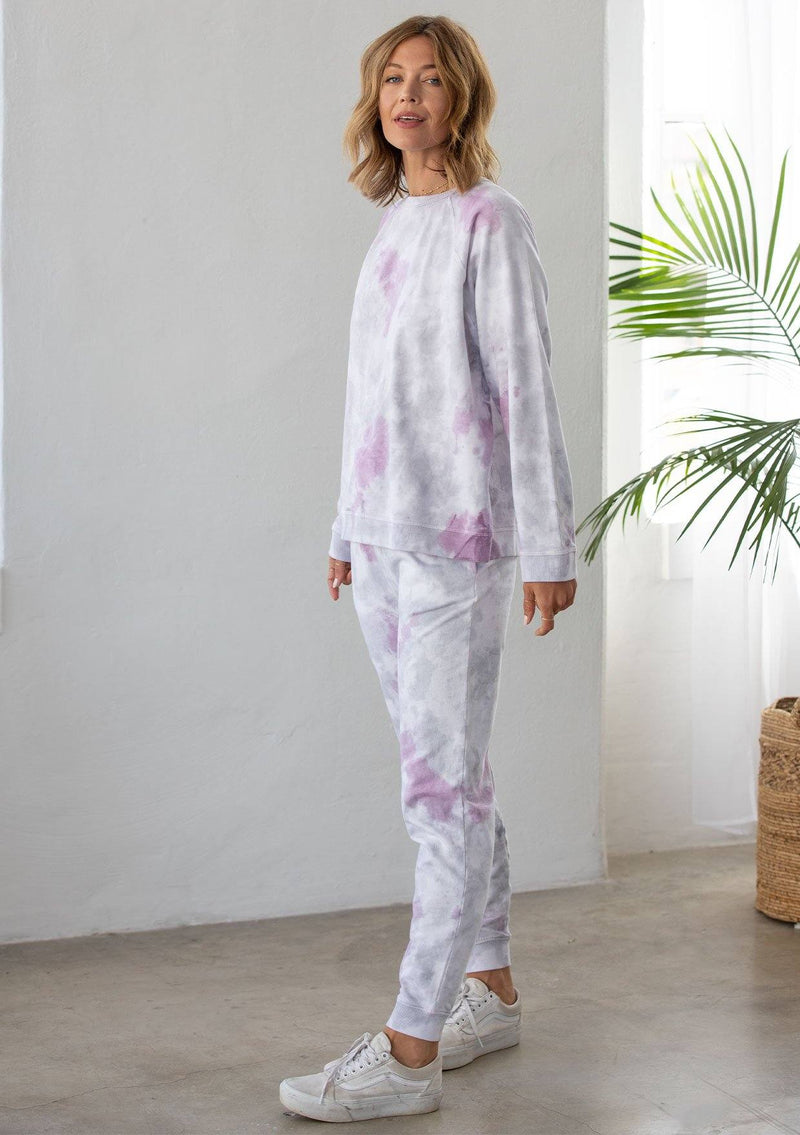 [Color: Lavender Combo] A woman standing outside wearing a classic cotton sweatshirt in a splatter tie dye wash. Featuring a crew neckline, long raglan sleeves, and a relaxed fit. 