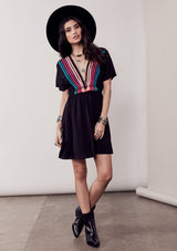 [Color: Black/Multi] A brunette model wearing a bohemian black dress with multicolored stripes and a tassel trim detail. Complete with a deep v-neckline, an elastic waist, and short sleeves. 