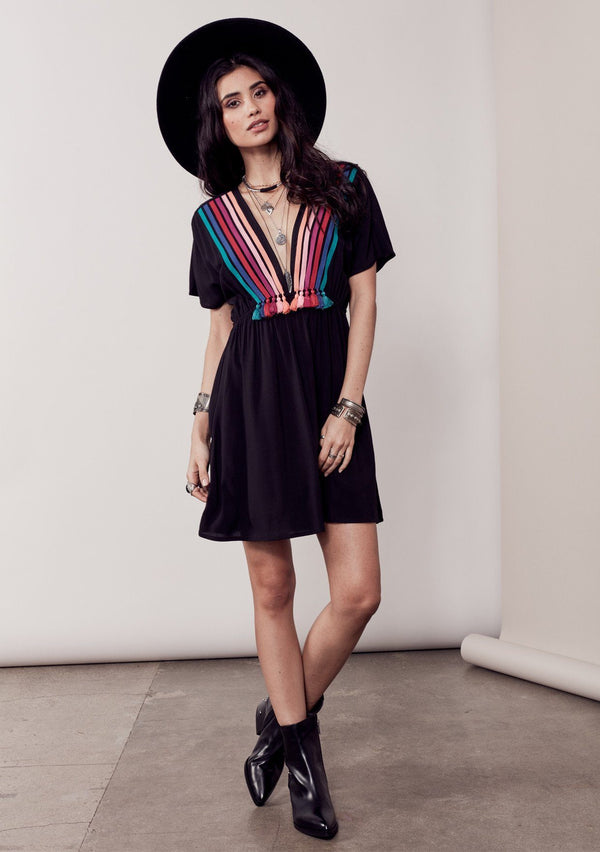 [Color: Black/Multi] A brunette model wearing a bohemian black dress with multicolored stripes and a tassel trim detail. Complete with a deep v-neckline, an elastic waist, and short sleeves. 
