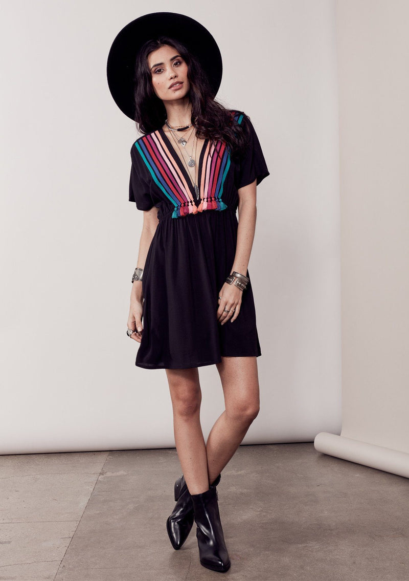 [Color: Black/Multi] A brunette model wearing a bohemian black dress with multicolored stripes and a tassel trim detail. Complete with a deep v-neckline, an elastic waist, and short sleeves. 