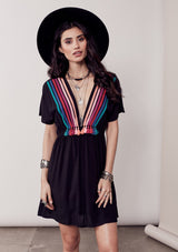 [Color: Black/Multi] A brunette model wearing a bohemian black dress with multicolored stripes and a tassel trim detail. Complete with a deep v-neckline, an elastic waist, and short sleeves. 
