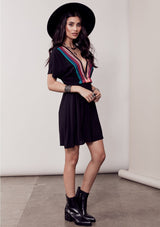 [Color: Black/Multi] A brunette model wearing a bohemian black dress with multicolored stripes and a tassel trim detail. Complete with a deep v-neckline, an elastic waist, and short sleeves. 