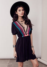 [Color: Black/Multi] A brunette model wearing a bohemian black dress with multicolored stripes and a tassel trim detail. Complete with a deep v-neckline, an elastic waist, and short sleeves. 