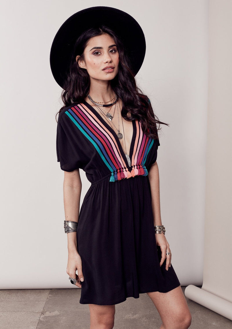 [Color: Black/Multi] A brunette model wearing a bohemian black dress with multicolored stripes and a tassel trim detail. Complete with a deep v-neckline, an elastic waist, and short sleeves. 