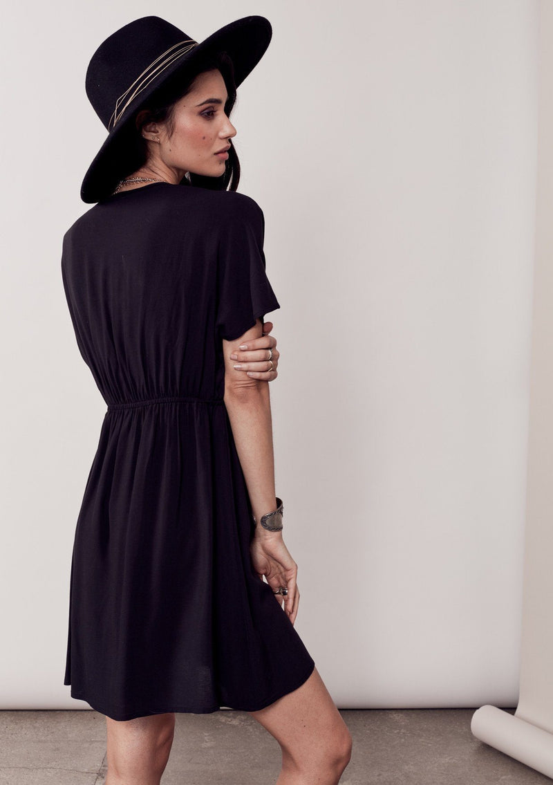 [Color: Black/Multi] A brunette model wearing a bohemian black dress with multicolored stripes and a tassel trim detail. Complete with a deep v-neckline, an elastic waist, and short sleeves. 