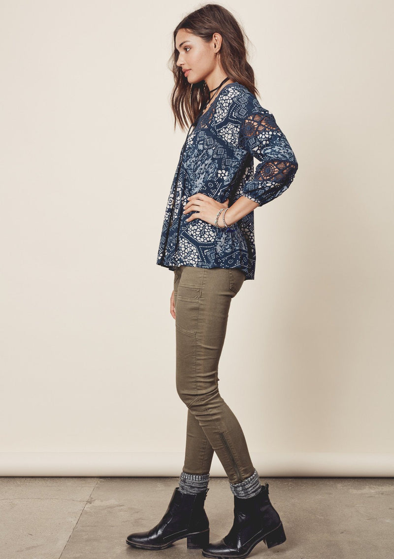 [Color: Teal/Vanilla] A side facing image of a brunette model wearing a teal  bohemian blouse with crochet lace trims. 