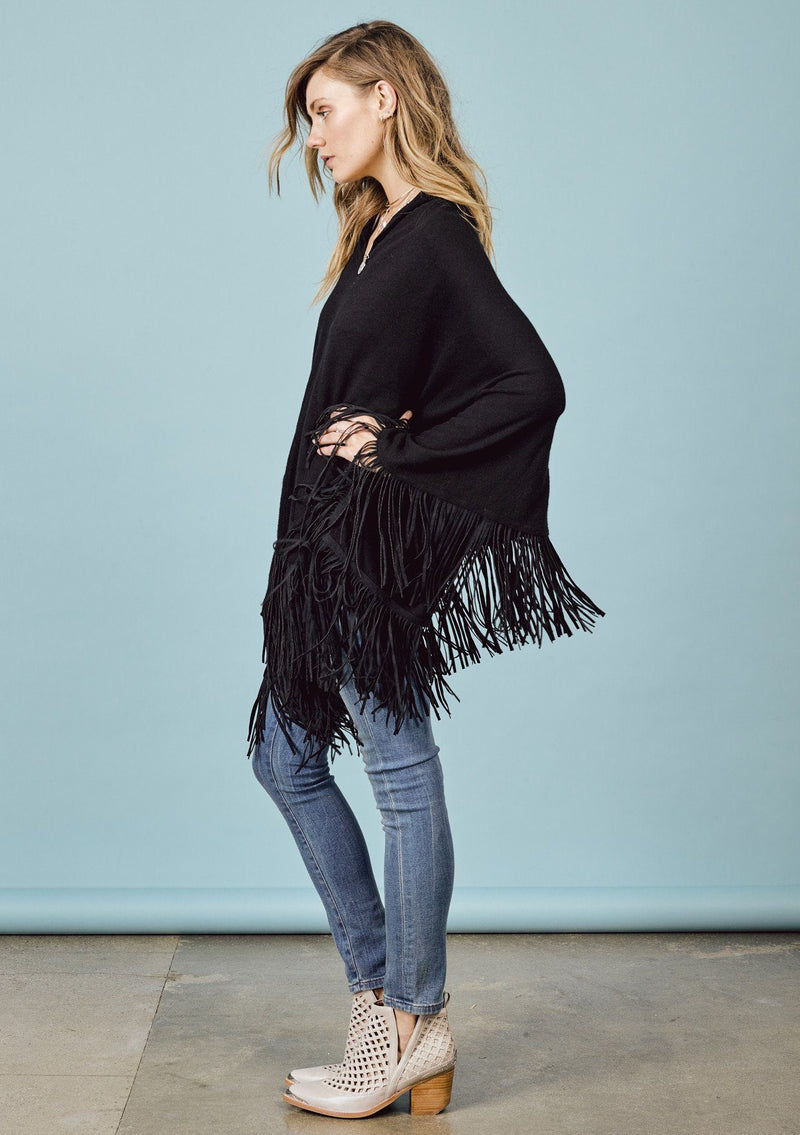 [Color: Black] A side facing image of a blonde model wearing a black suede fringed poncho.  