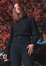 [Color: Jet Black] A blonde model wearing a black long sleeve Victorian style top with eyelet, mixed trim, mocked neckline, and smocked cuffs. 