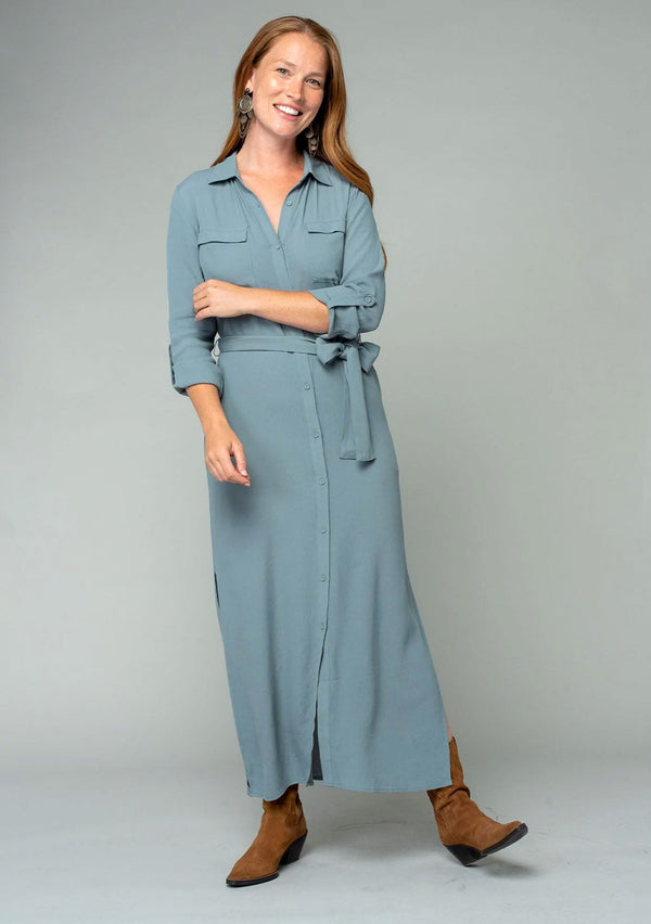 [Color: Balsam Green] A front facing image of a red headed model wearing a relaxed fit light green maxi shirt dress made from a lightweight crepe. With long rolled sleeves, a button tab sleeve closure, two front pockets, side pockets at the hip, and a self tie waist belt.