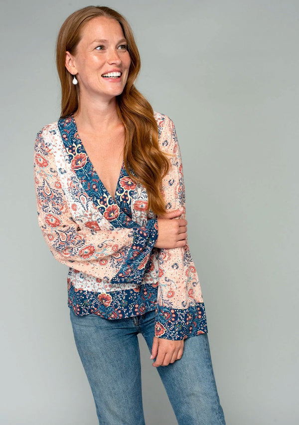 [Color: Ivory/Coral] A side facing image of a red headed model wearing an ivory white and coral pink mixed floral bohemian top. With a plunging surplice v neckline, long bell sleeves, and an elastic peplum waist.