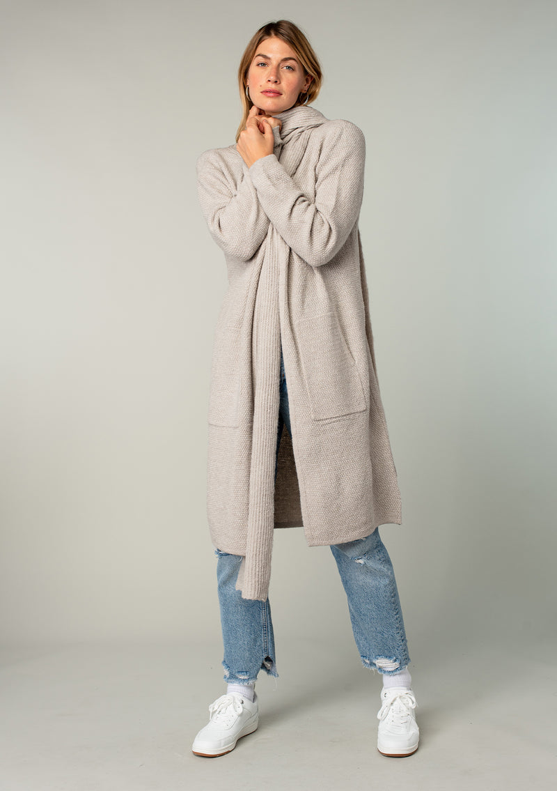 [Color: Mushroom] A model wearing a cozy taupe long shawl cardigan. With long sleeves, side pockets, an open front, and attached scarf detail.